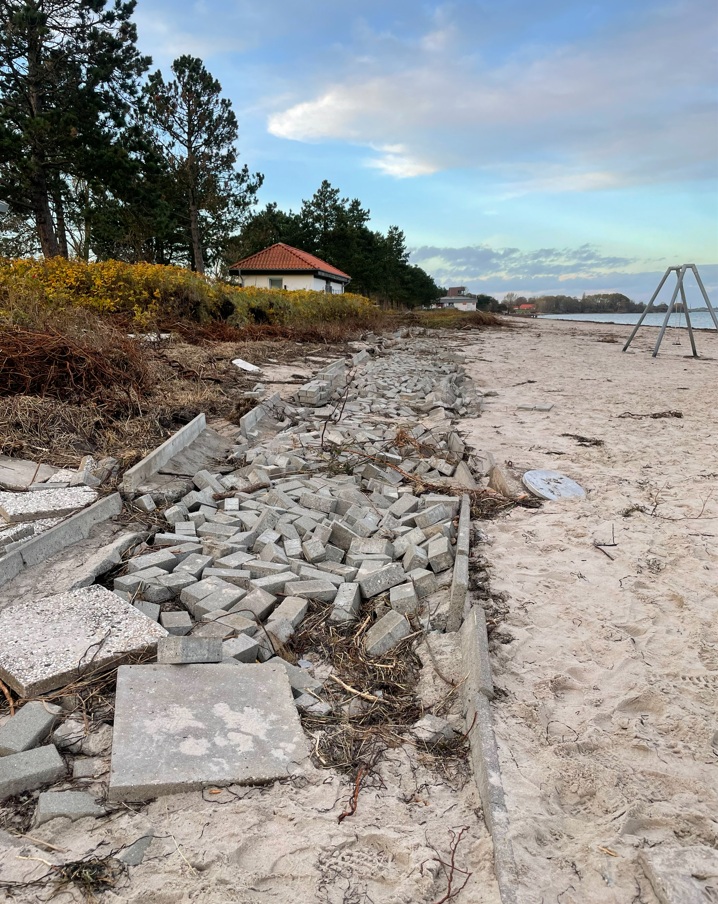 Bild von einem zerstörten Pflasterweg mit eingespülten Pflanzenresten, rechts davon Strand und weiter hinten noch hinter einem Schaukelgerüst gerade so erkennbar die Küstenlinie.